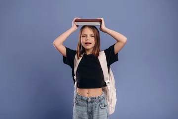 Young girl with brown hair wearing a black shirt and denim shorts holds a book above her head, smiling against a solid blue background © standret