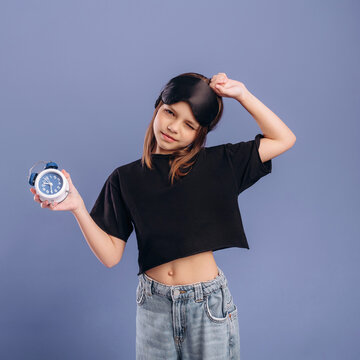 Young girl wearing sleep mask and holding alarm clock poses playfully in front of a solid purple background, showcasing a casual and relaxed style