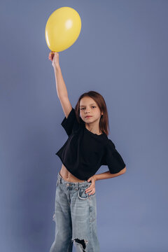 Young girl with brown hair wearing black crop top and ripped jeans holds a yellow balloon high against a solid blue background in a playful pose