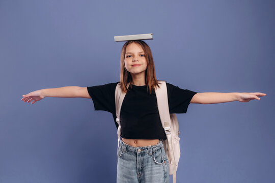 Young girl balancing a book on her head with arms outstretched, wearing a black t-shirt and backpack against a solid blue background