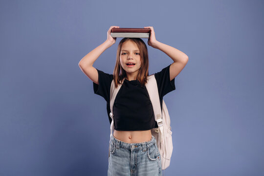 Young girl with brown hair wearing a black shirt and denim shorts holds a book above her head, smiling against a solid blue background