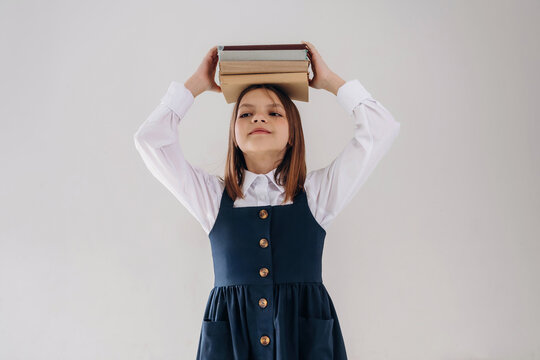 Young girl with brown hair in a blue dress balances books on her head while standing against a plain white background, showcasing confidence and poise in a playful manner