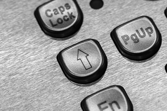 Macro black-and-white close-up of metal keyboard keys showing an up arrow, Caps Lock and PgUp buttons. High-contrast texture emphasizes technology, control, typing and industrial design concepts