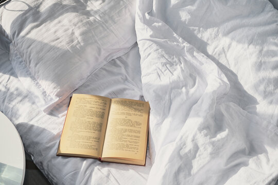 Open book resting on rumpled white linen sheets and pillow in morning light