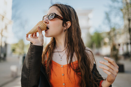 A young woman wearing sunglasses eats an ice cream cone while standing on a city street. The casual summer portrait shows relaxed style and enjoyment outdoors.
