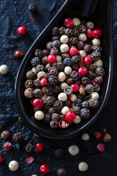 Variety of colorful peppercorns in a black spoon on dark textured surface.
