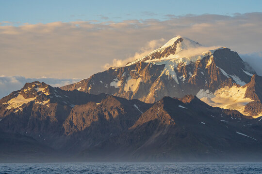 View of snow-capped mountain peaks near Grytviken, illuminated by golden hour light, with rugged slopes and calm sea, Grytviken, South Georgia and the South Sandwich Islands.