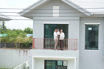 Obraz premium Young parents holding their child stand at the wooden railing admiring the neighborhood view standing on the terrace of a newly built suburban home in warm natural light