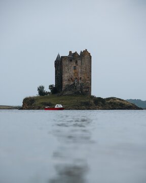 View of Castle Stalker on a small island in a loch with a red boat nearby under an overcast sky in Appin, Scotland, United Kingdom.