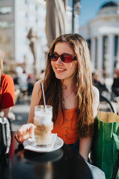 A young woman wearing sunglasses smiles while holding an iced coffee at an outdoor cafe. She enjoys a sunny cafe moment in casual summer clothing with a shopping bag nearby.
