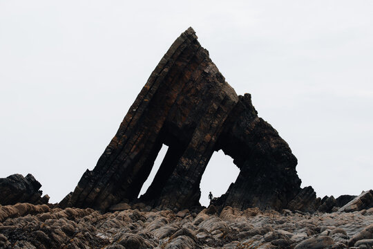 View of Blackchurch Rock with a person standing beneath the dramatic, jagged rock formation on a rocky beach in Westward Ho!, United Kingdom.