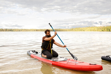 Obraz premium Three quarter front view of a bearded man kneeling and paddling with a blue shaft paddle on a red inflatable SUP board, calm lake water, distant green forest under cloudy sky, daytime horizontal shot