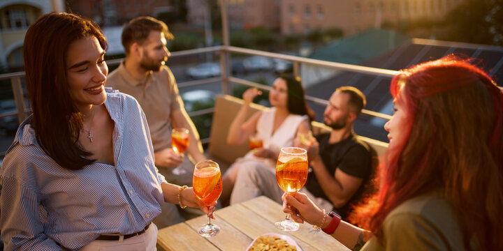 Two women share orange Aperol Spritz cocktails at rooftop gathering with relaxed happiness. Female friendship, summer leisure and social lifestyle concept.