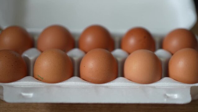 A close-up shot of ten brown chicken eggs arranged neatly in two rows inside an open white styrofoam carton box on a wooden surface.