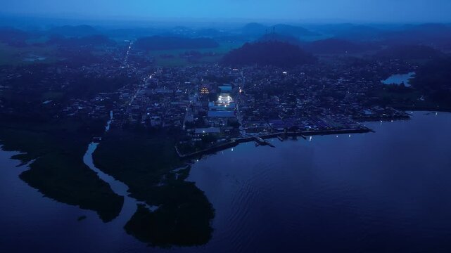 Cinematic drone view of an illuminated basketball court during a community event in Buhi, Camarines Sur, Philippines. Seen from far above and across the town at blue hour, the intense court lighting c