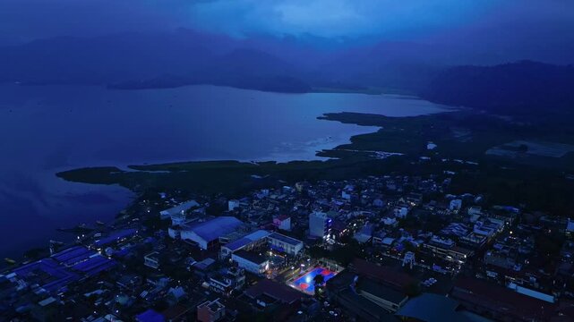 Cinematic aerial footage of a brightly illuminated basketball court during a community event in Buhi, Camarines Sur, Philippines. Captured from a distant perspective at blue hour, the sports court app