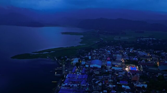 Cinematic drone view of an illuminated basketball court during a community event in Buhi, Camarines Sur, Philippines. Seen from far above and across the town at blue hour, the intense court lighting c