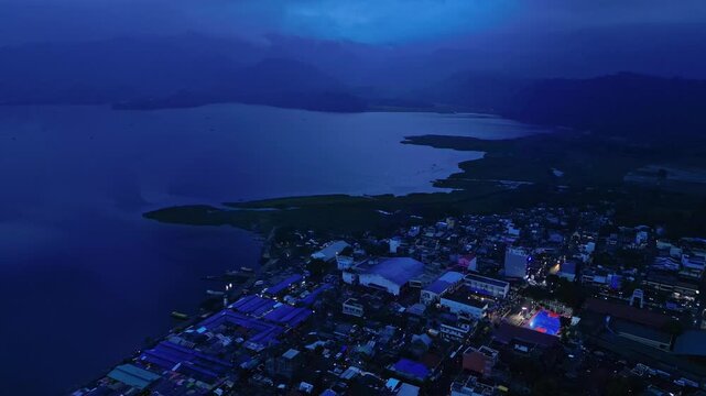 Wide aerial view of Buhi town at dusk with a brightly illuminated basketball court standing out against the surrounding landscape. The court is hosting a local community event and appears as a distinc