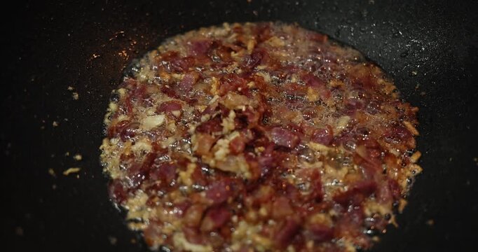Crispy bacon pieces fry in bubbling fat inside black frying pan. Chopped pork browns evenly developing crunchy texture at cooking process in kitchen