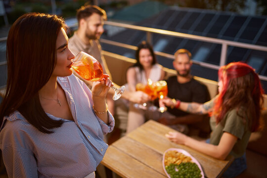 Young woman sips citrus cocktail beside rooftop friends with relaxed pleasure. Social lifestyle, refreshing drinks and summer leisure concept.