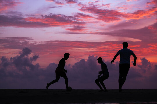 View of three people playing soccer on the beach in silhouette against a vibrant sunset sky in Majorda, Goa, India.