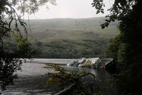 View of a partially submerged boat in the calm waters of Loch Lochy, framed by lush green trees and rolling hills in Blairgowrie, Scotland, United Kingdom.