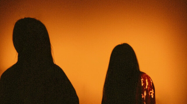Two Long Haired Shadowy Figures in a Grainy Orange Glow
