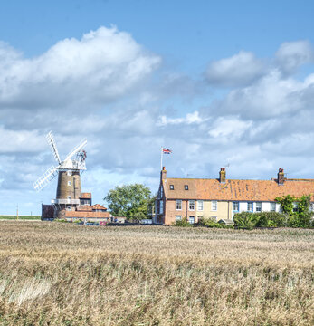 View of Cley Windmill standing beside a row of historic brick houses in a grassy field under a blue sky with white clouds in North Norfolk District, England, United Kingdom.