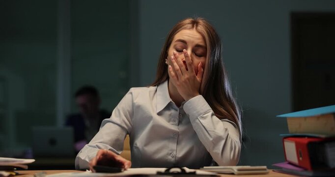Tired office woman checks late time on smartphone at dark desk. Exhausted employee yawns struggling to stay awake during overtime work slow motion