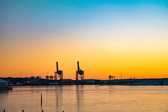 View of port cranes silhouetted against a vibrant sunset sky over a calm, icy harbor in Oslo, Oslo, Norway.