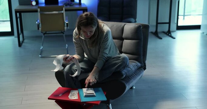 Woman sits in armchair with stack of receipts and calculator on laps. Careful person flips through pages calculating expenses with focused attention