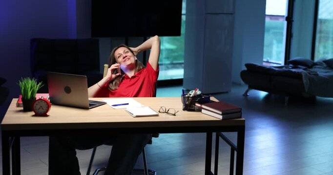 Relaxed office woman sits at modern desk with laptop and phone. Businesswoman talks on mobile leaning back during casual afternoon work break