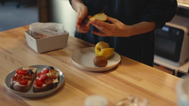 Close-up of woman's hands cutting core from Golden apple quarter with knife. Preparing fresh fruit with kiwi and tomato sandwiches on wooden table in home kitchen. Healthy snack preparation concept.