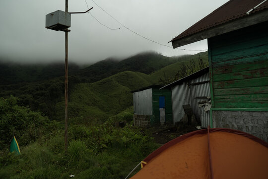View of an orange tent set up near rustic wooden and corrugated metal buildings with lush green rolling hills and misty mountains in the background in Kohima, Nagaland, India.