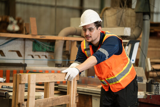 A young male worker wearing hard hat, safety glasses, and protective vest inspects a wooden pallet frame inside a wood factory. Carpenter working at the station