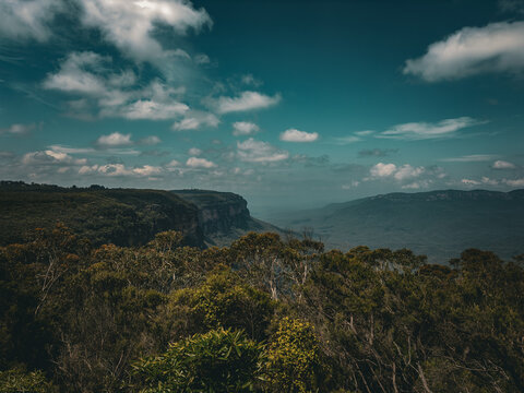 View of the vast Blue Mountains escarpment with dense eucalyptus forests, rugged sandstone cliffs, and a dramatic cloudy sky in Blue Mountains City Council, New South Wales, Australia.