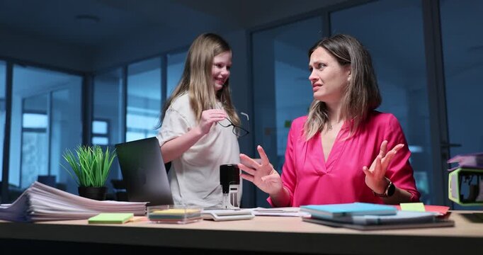 Girl approaches mother sitting at office desk during work. Busy woman smiles at child responding warmly while girl interrupts regular daily tasks