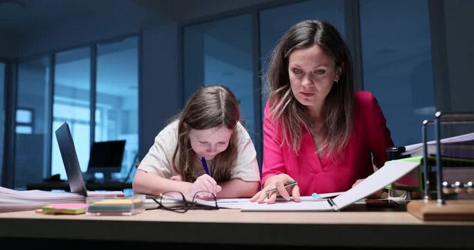 Working mother sits at office desk near daughter with drawing paper. Woman writes notes while preteen girl colors picture staying close during work