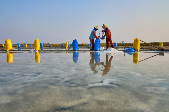 View of salt workers in conical hats harvesting salt in shallow pans with colorful bags and reflections in Kaliori, Central Java, Indonesia.