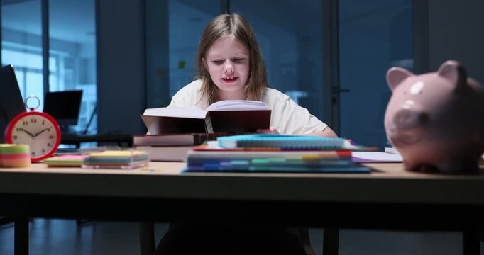 Focused girl reads thick textbook at desk with stacked books. Junior student studies difficult material concentrating on page with serious expression