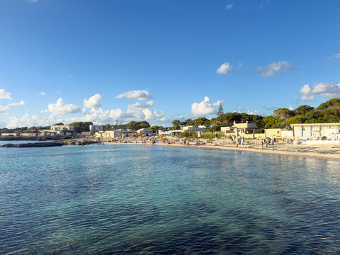 View of the Island of Favignana beach with clear turquoise water, sunbathers on the sand, and coastal buildings under a bright blue sky in Favignana, Sicily, Italy.