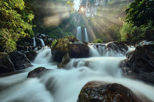 View of a cascading waterfall flowing over dark rocks with lush green forest foliage and sun rays filtering through the trees in Magelang Regency, Central Java, Indonesia.