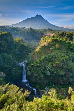 View of Mount Merapi towering over a lush tropical valley with a cascading waterfall surrounded by dense forest and vibrant green foliage in Magelang, Central Java, Indonesia.