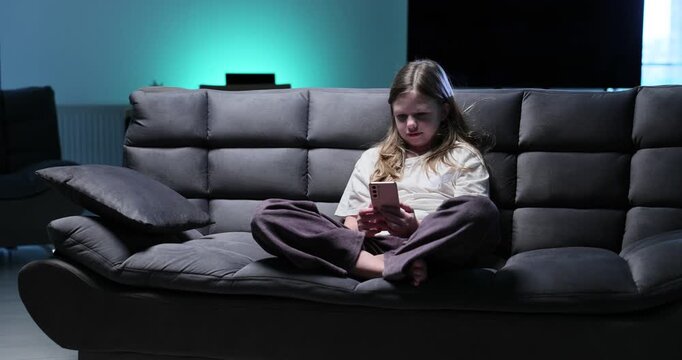 Sad lonely preteen girl watches short videos on smartphone sitting on sofa. Pensive schoolgirl addicted to internet scrolls social networks at home