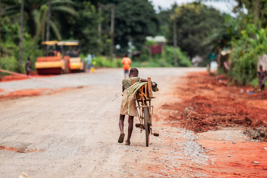 View of a young boy walking along a dusty road carrying a bicycle loaded with palm fronds, with construction machinery visible in the background, Port Harcourt, Rivers, Nigeria.