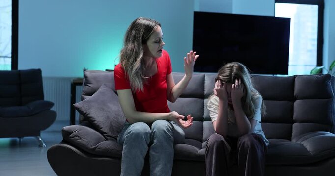 Strict mother scolds upset little girl clutching head as sits on sofa at home. Woman expresses disappointment of daughter behavior in living room
