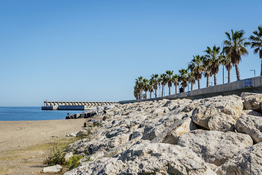 View of a rocky breakwater along the coast with a row of palm trees lining the promenade under a clear blue sky in Malaga, Andalusia, Spain.