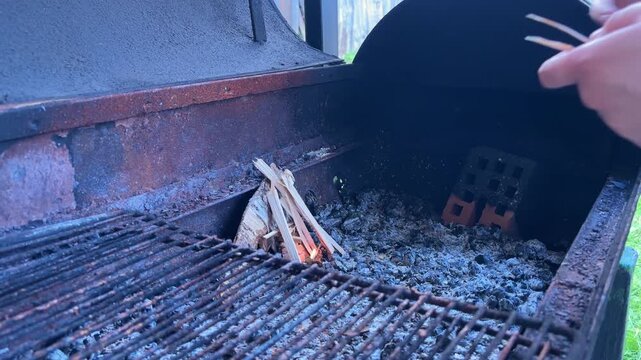 Hands arrange kindling and light a small fire inside a charcoal grill, outdoor barbecue preparation process.