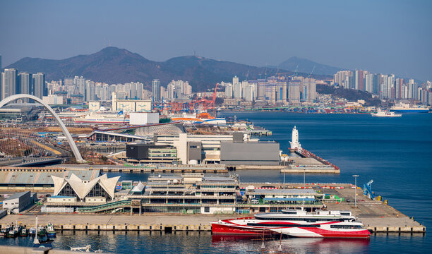 View of the busy port of Busan with modern ferry boats, industrial infrastructure, and a sprawling cityscape set against coastal mountains under a clear blue sky in Busan, Busan, South Korea.