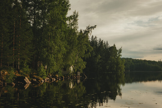 View of a serene lake bordered by a dense forest of pine and birch trees with a small cabin nestled along the shoreline under a soft, overcast sky in Hameenlinna, Kanta-Hame, Finland.